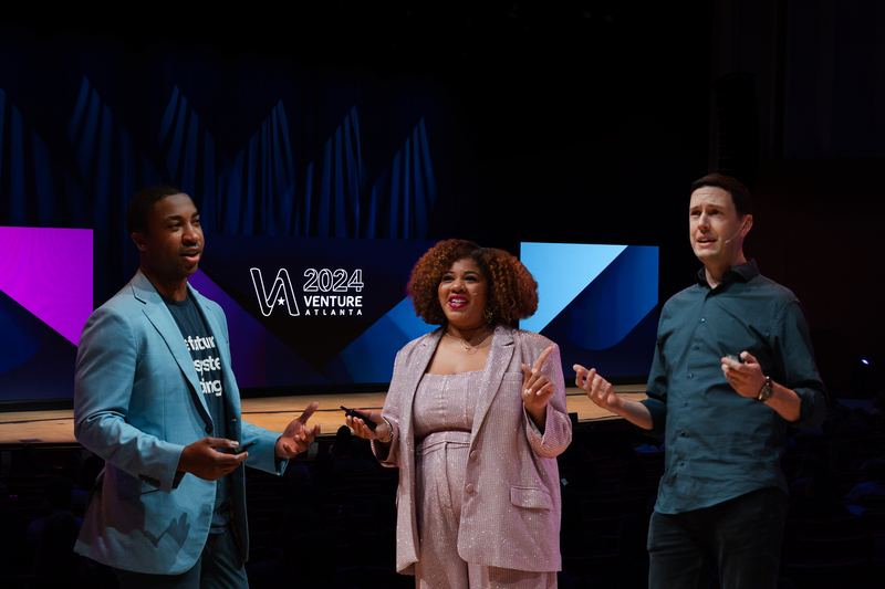 Two founders stand in front of a stage with an investor in a conference style room